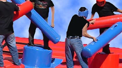 Four people wearing helmets joust with padded sticks on an inflatable red and blue platform under a blue sky.