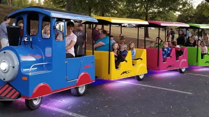 Colorful miniature train with blue, yellow, pink, and green cars carrying children and adults in a park setting.