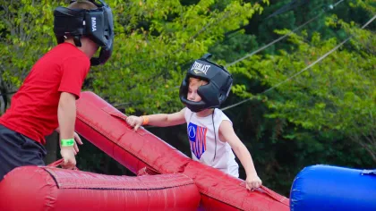 Two children wearing protective headgear joust with large padded sticks during an outdoor play activity.