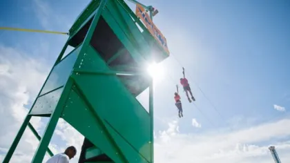 Two people ziplining from a green tower structure on a sunny day with clear blue sky.