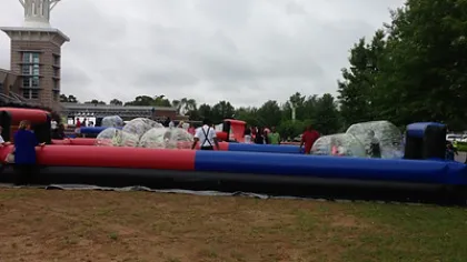 Outdoor bubble soccer game with players inside inflatable bubbles on a grassy field under cloudy sky.