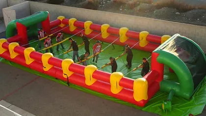 People playing inflatable human foosball game with red and yellow barriers on green field outdoors.