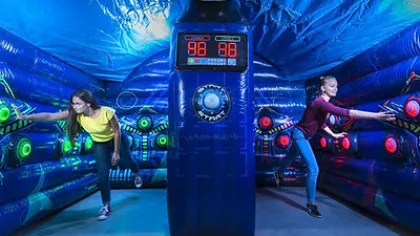 Two girls playing laser tag in a dark, neon-lit inflatable arena with glowing targets and digital score display.