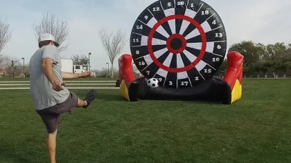 Man kicking a soccer ball towards a large inflatable dartboard target on a grassy field outdoors.