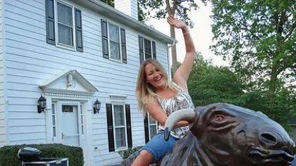 Happy woman riding a mechanical bull outside a white house during daytime, waving and smiling.