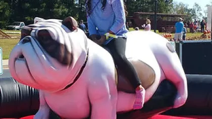 Young girl riding a mechanical bulldog ride at an outdoor event on a sunny day.