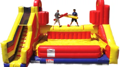 Two people jousting on a large red and yellow inflatable obstacle course with a slide.