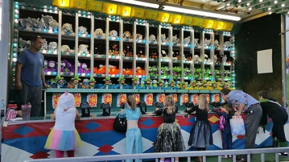 Children and adults playing a carnival shooting game with stuffed animal prizes displayed on shelves.