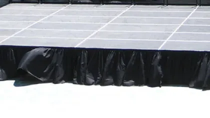 Empty outdoor portable stage with black skirt and stairs, set up in a sunlit open area.