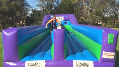 Man in life jacket playing on a colorful purple and blue inflatable water obstacle course outdoors.