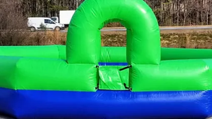 Large green and blue inflatable structure with an archway, set outdoors on concrete with trees and vehicles in the background