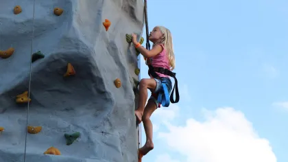 Young girl in pink shirt climbing an artificial rock wall outdoors with safety harness against blue sky.