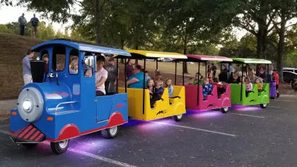 Colorful miniature train with blue, yellow, pink, and green cars carrying children and adults in a park setting.