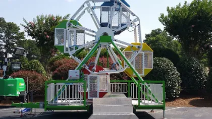 Small colorful ferris wheel ride set up outdoors with green and white fencing under clear skies