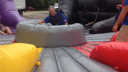 Man in blue shirt and harness playing on inflatable obstacle course with colorful barriers outdoors.
