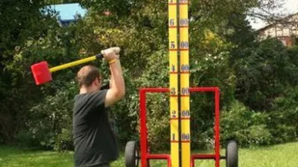 Man striking a carnival strength tester game with a large red mallet outdoors on grass near trees.