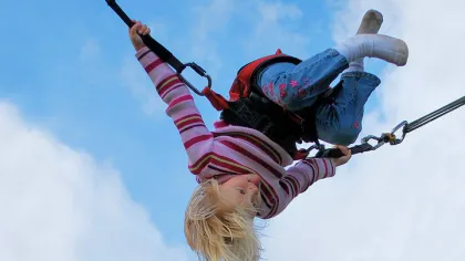 Young child with blonde hair swinging upside down on a harness against a blue sky.