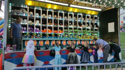 Children and adults playing a carnival game with stuffed animal prizes in numbered shelves at a fair booth.