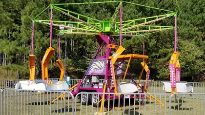 Colorful empty carnival swing ride with yellow, pink, and green accents surrounded by metal fencing on grass.