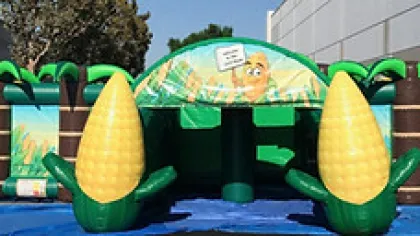 Inflatable bounce house with palm trees and giant corn cobs on the sides in an outdoor setting.