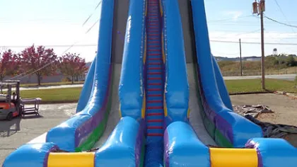 Large colorful inflatable slide with dual lanes and central climbing stairs outdoors on a sunny day.