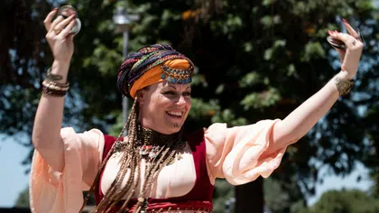 Smiling woman in colorful traditional attire dancing outdoors with raised arms and accessories under sunlight.