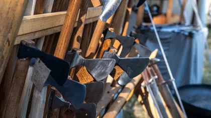 Rack of various axes with wooden handles displayed outdoors in daylight at a market or store.