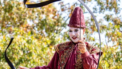 Performing circus clown in vintage costume balancing on a rolling board outdoors with flowing ribbon wand.