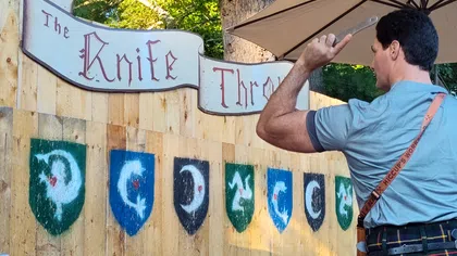 Man aiming a knife at a wooden target board labeled The Knife Throw with shield symbols outdoors under an umbrella.