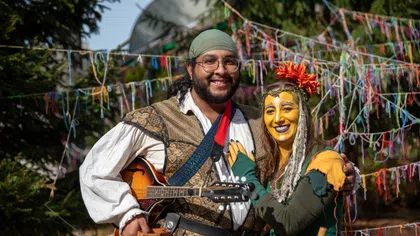 Smiling couple in colorful renaissance costumes with guitar and flower headpiece at outdoor festival.
