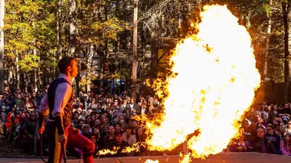 Fire performer breathing a large flame on stage during an outdoor show with a crowd watching.