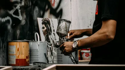 Man holding paint sprayer near paint cans and a framed art portrait in a workshop with graffiti background.