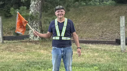 Man wearing reflective safety vest and cap waving an orange flag in a grassy outdoor area near trees.