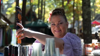 Smiling woman in purple blouse pouring a seasonal beer at an outdoor autumn festival with plastic cups and trees in background