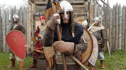Medieval warriors in armor with shields and weapons standing outside a wooden fort gate on grass.