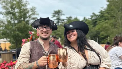 Smiling couple in pirate costumes holding copper mugs and sitting outdoors by flowers and brick wall.