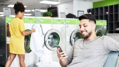 Smiling man using smartphone while woman loads laundry into washing machine at laundromat with green accents