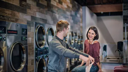 Young man and woman chatting in a modern laundromat with stacked washers and dryers along the wall.