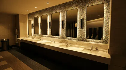 Modern public restroom with six illuminated mirrors and marble countertop sinks in warm lighting.