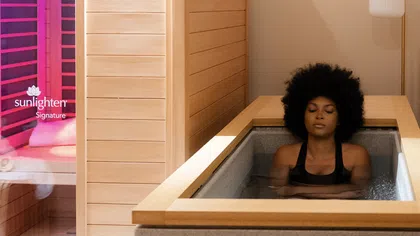 Woman relaxing in a wooden spa bath tub next to a lighted sauna cabin, showing wellness and calmness.