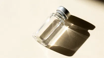 Clear glass vial with metal cap casting a shadow on a beige surface in bright natural light.
