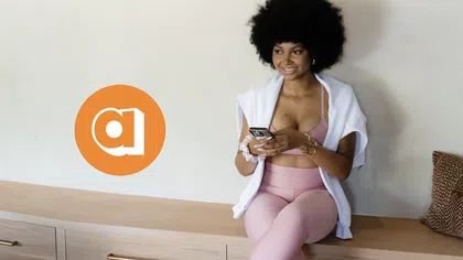 Smiling woman in pink activewear sitting on wooden bench using smartphone with orange app logo on wall.