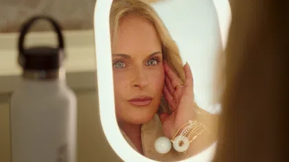 Woman with blonde hair and blue eyes adjusts her earring looking into an illuminated oval mirror.