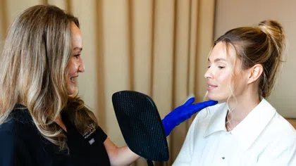 Cosmetologist with blue gloves examines woman's chin while she holds a black hand mirror during consultation.