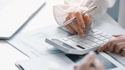 Person using calculator and analyzing financial documents next to laptop on white desk
