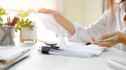 Person reviewing documents at desk with pen in hand in a bright, organized workspace.