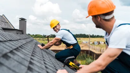 Two construction workers in orange and yellow helmets installing shingles on a rooftop under a cloudy sky.