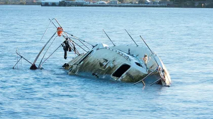 Partially submerged boat sinking in calm water near shoreline with distant trees and buildings.