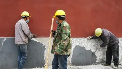 Three construction workers wearing yellow helmets and masks repair and clean a wall with peeling paint.