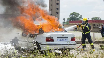 Firefighter extinguishing flames on a white car fully engulfed in fire with thick black smoke outdoors.